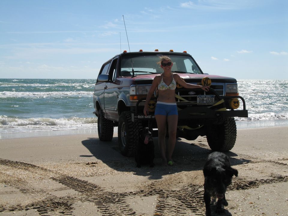 Lifted red Ford Bronco with brush guard parked on a beach beside the surf