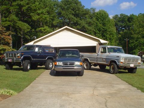 1984 Ford Bronco - A blue 1984 Ford Bronco is parked in a driveway alongside two other trucks, with a garage in the background.