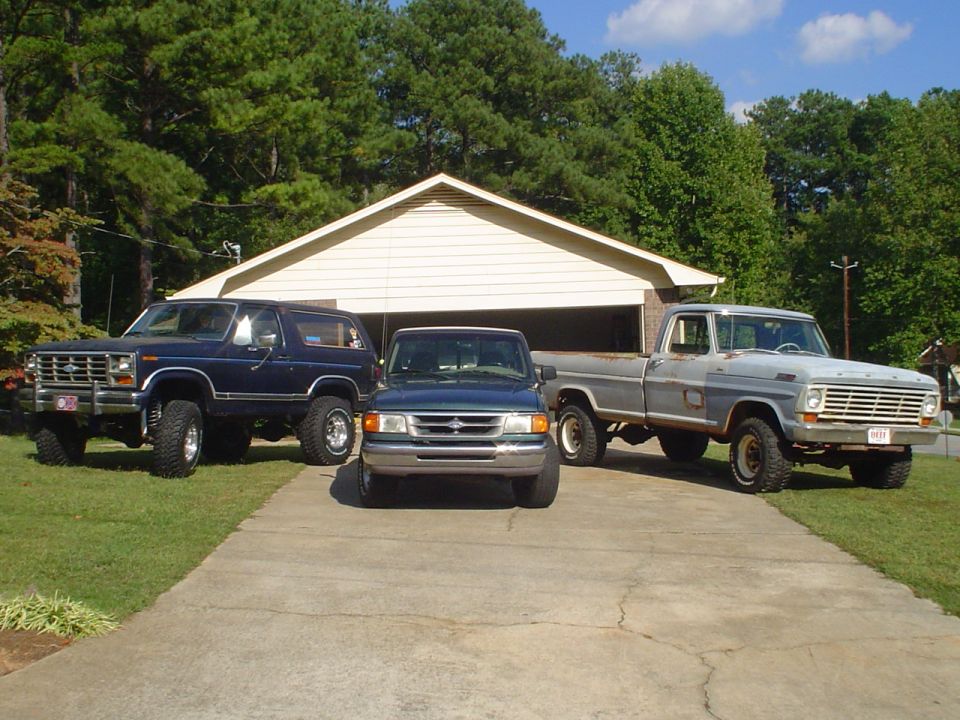 Blue 1984 Ford Bronco parked in front of a garage with two trucks