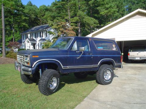 1984 Ford Bronco - Lifted blue Ford Bronco sits on the lawn next to a garage containing another vehicle.