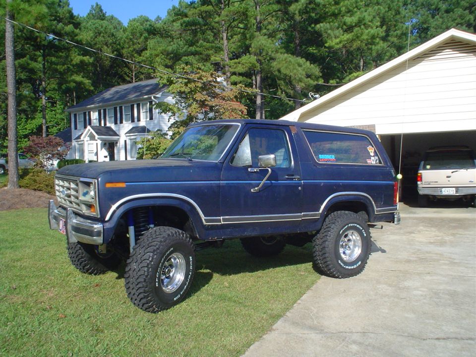 Blue lifted Ford Bronco parked on grass, another SUV in garage behind it