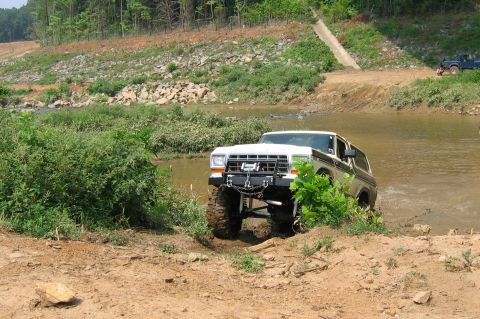 1992 Ford Mustang - A lifted Ford Bronco navigates a muddy water crossing, with a blue pickup truck in the distance.