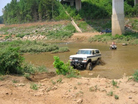 1992 Ford Mustang - A white Ford Bronco navigates a muddy waterway under a bridge, with other vehicles visible in the background.