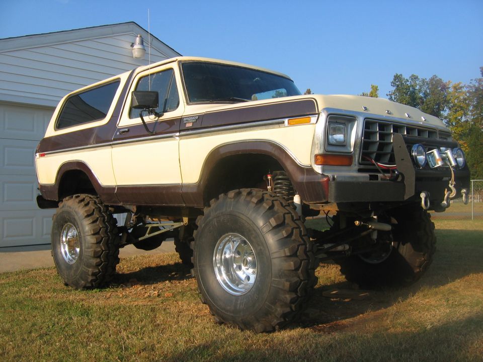Lifted two-tone beige and brown Ford Bronco with large off-road tires parked on grass in front of a garage