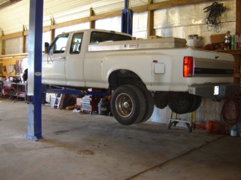 1992 Ford Bronco - A white dually pickup truck is elevated on a hydraulic lift inside a workshop, showing its rear underside and dual rear wheels.