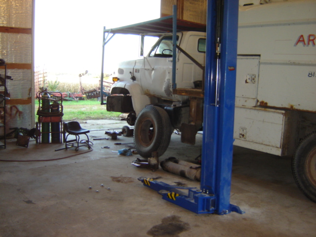 White truck with its front wheel off, on a lift in a garage. Tools and parts are scattered around the floor