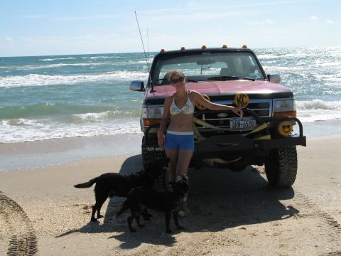 1995 Ford Bronco - Lifted pink and black Ford Bronco parked at the surf line on a beach, photographed head-on with a tall CB whip antenna, roof-mounted cab li…