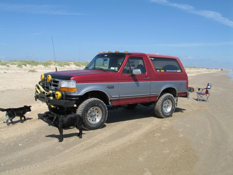 1995 Ford Bronco - Lifted two-tone red and silver Ford Bronco parked on a wide sand beach, wearing a custom front bumper with a yellow-drum winch, dual drivin…