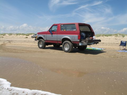 1995 Ford Bronco - Red-over-silver two-tone full-size Ford Bronco parked on wet beach sand, shot from the rear three-quarter with the rear glass raised and ta…