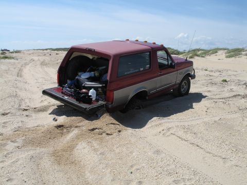 1995 Ford Bronco - Rear three-quarter view of a two-tone burgundy-over-tan full-size Ford Bronco parked on sandy beach dunes, tailgate down and rear glass hat…