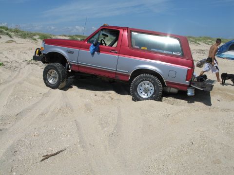 1995 Ford Bronco - Side view of a lifted two-tone red and silver Ford Bronco flexing over a sand dune at the beach, riding on Ventura mud-terrain tires and po…