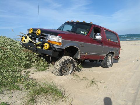 1995 Ford Bronco - Two-tone dark red over silver full-size Ford Bronco flexing its front suspension as it climbs a beach dune, shot from the front three-quart…