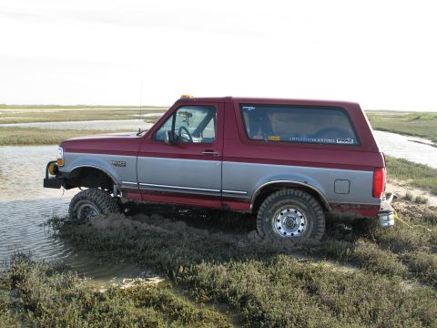 1995 Ford Bronco - A maroon-and-silver two-tone full-size Ford Bronco sunk to its axles in marsh mud, driver-side profile riding on mud tires with a tubular f…