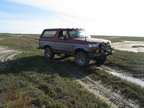 1995 Ford Bronco - Lifted two-tone maroon-over-silver Ford Bronco parked on muddy tidal-flat terrain, with cab roof clearance lights, mud-terrain tires on ste…