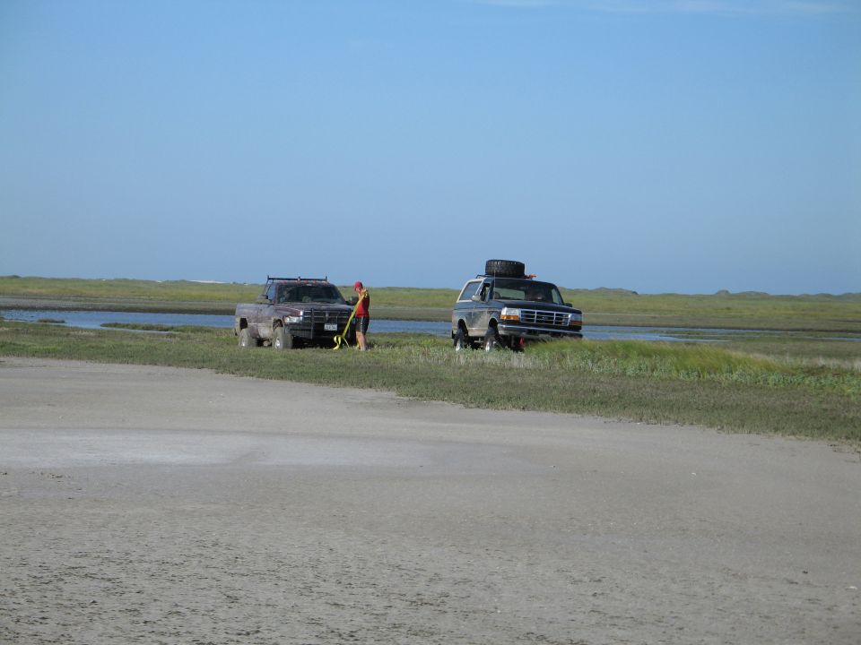 Dark pickup tow-strapped to silver Ford Bronco on coastal marsh flat