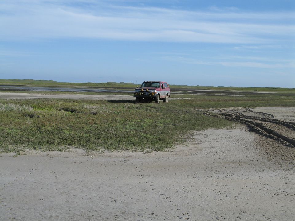Red-and-white Ford Bronco parked on open sandy flats under a blue sky, distant view