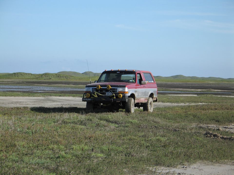 Lifted red 1990s Ford Bronco with brush guard parked on grassy wetland flats