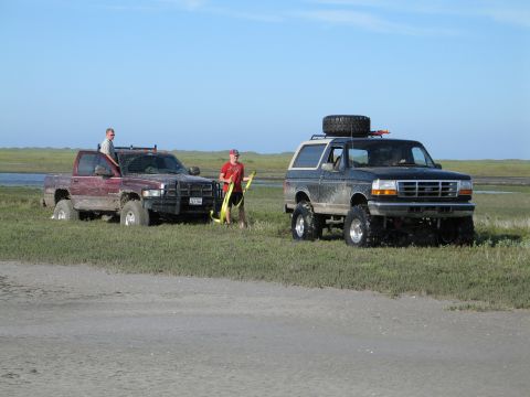 1995 Ford Bronco - Recovery on a marshy flat near dunes: a person holds a yellow tow strap between a mud-splattered maroon Dodge Ram pickup with a grille guar…