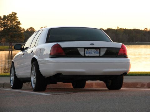 2003 Ford Crown Victoria - Three-quarter rear view of a white Ford Crown Victoria parked at a lakeside lot at dusk, showing a blacked-out trunk panel between the tail…