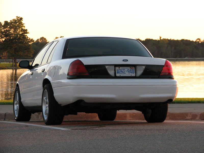 Rear three-quarter view of white Crown Victoria with blacked-out trunk panel by a lake