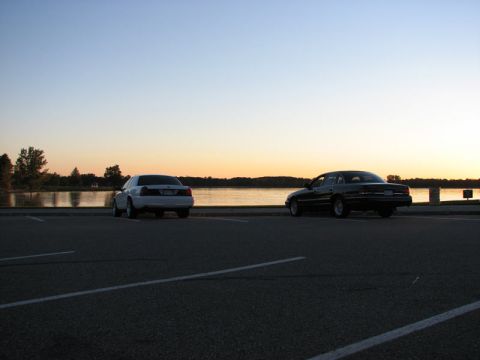2003 Ford Crown Victoria - Two full-size sedans, one white, one black, parked nose-out along a lakeside lot at sunset, silhouetted against orange sky and calm water.…