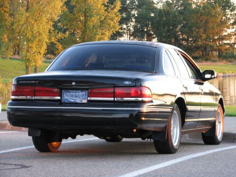 2003 Ford Crown Victoria - A black Ford Crown Victoria LX sedan seen from the rear three-quarter, with full-width red tail lights, chrome trim and a Michigan plate. I…