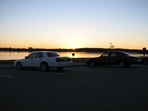 2003 Ford Crown Victoria - A white Ford Crown Victoria full-size sedan on five-spoke alloy wheels sits parked in a lakeside lot at sunset, facing away from a black Cr…