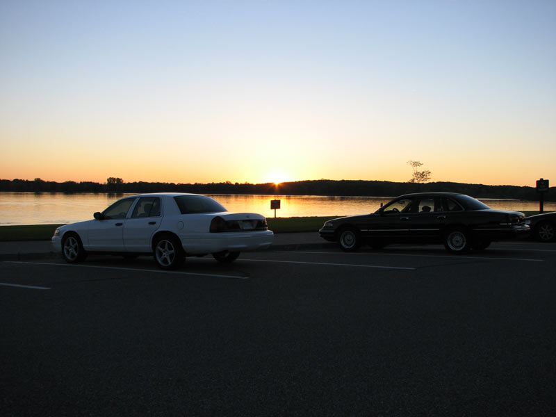 White Crown Victoria sedan parked beside a black one at a lakeside lot at sunset