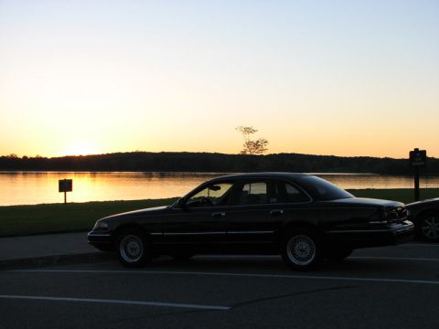 2003 Ford Crown Victoria - Dark full-size Ford sedan photographed in near-silhouette from the driver's side, parked in an empty lakeside lot at sunset with the sun se…