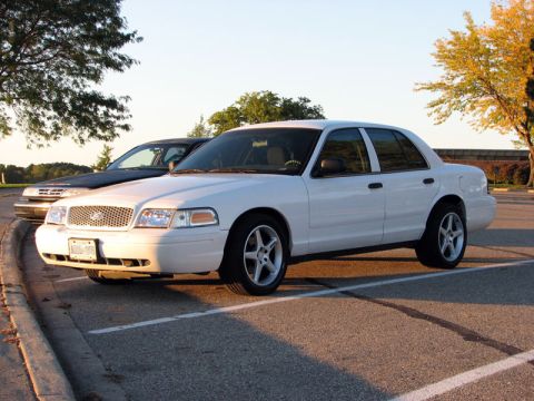 2003 Ford Crown Victoria - White Ford Crown Victoria sedan photographed from a front three-quarter angle in an empty parking lot at golden hour, with aftermarket silv…