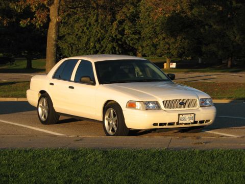 2003 Ford Crown Victoria - White Ford Crown Victoria sedan in a three-quarter front view, parked across parking-lot spaces in low evening sunlight with trees behind.…