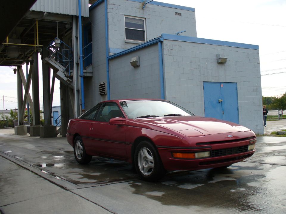 Red Ford Probe coupe, front 3/4 view, parked by an industrial building.