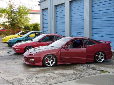 1991 Ford Probe - Row of Acura Integra coupes parked at an angle in front of blue roll-up shop doors, led by a lowered red car with a full body kit, wide chr…