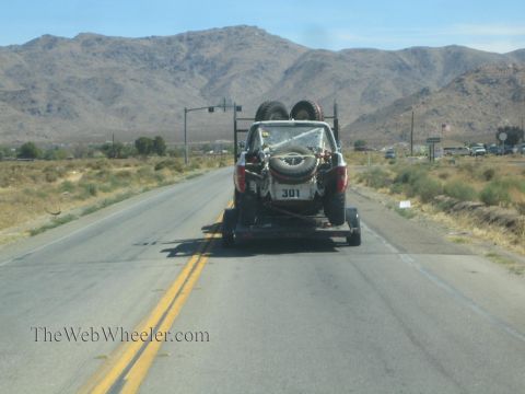 1990 Ford Bronco - View through a windshield of an off-road race car on a flatbed trailer being towed along a two-lane desert highway.