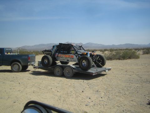 1990 Ford Bronco - sponsor-decaled off-road race buggy sits strapped to a tandem-axle flatbed trailer hitched to a pickup in a desert staging area.