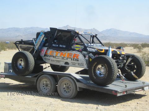 1990 Ford Bronco - black Letner Racing off-road race buggy with flame graphics and spare tires sits chained to a flatbed trailer in a desert setting.