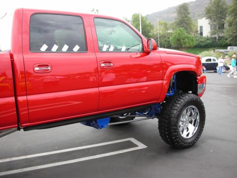 1992 Ford Crown Victoria - A red pickup truck with custom suspension is parked in a lot, with other vehicles and people in the background.