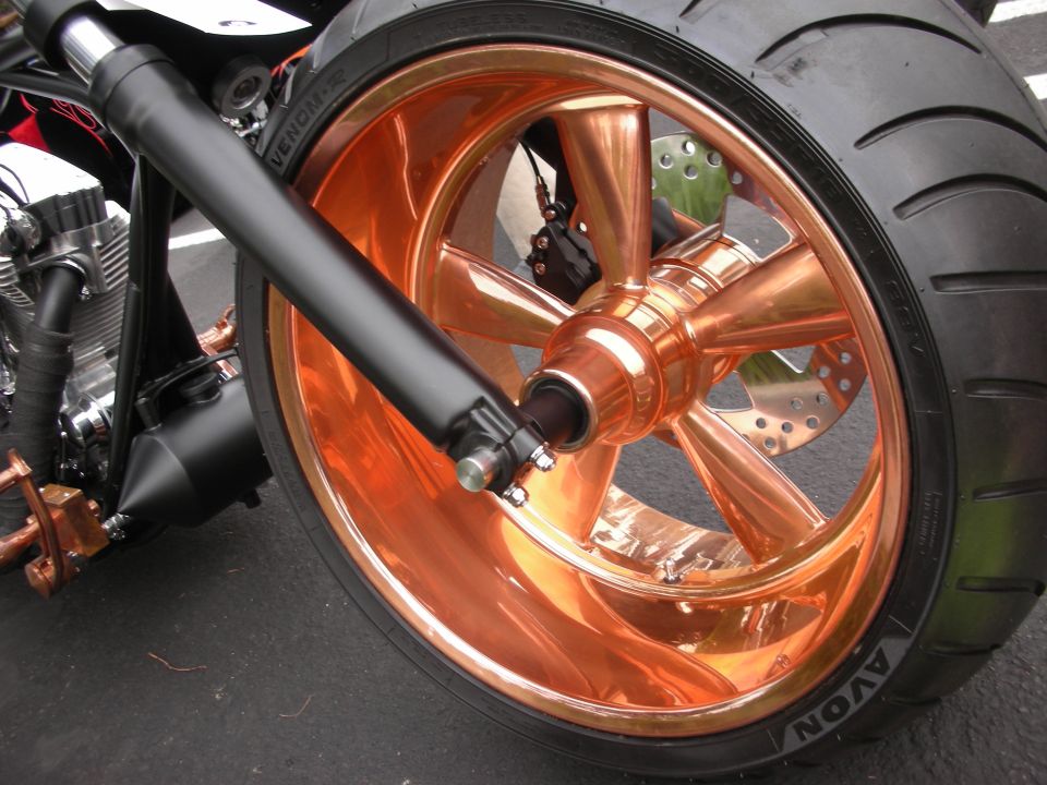 Close-up of a motorcycle's front wheel with a shiny copper-finished rim and spokes. Black front fork is visible