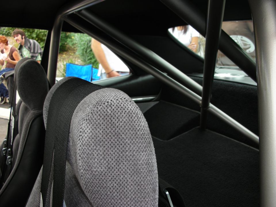 Interior view of a car showing grey racing seats, black seatbelts, and a metal roll cage. People are visible through the rear window
