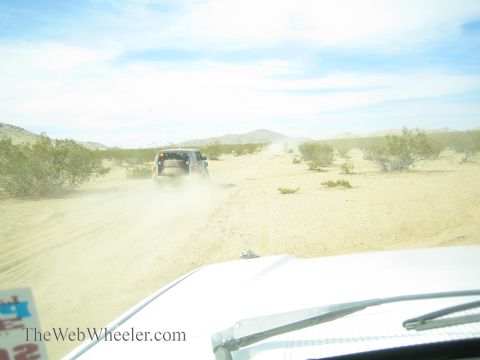 1990 Ford Bronco - off-road vehicle raises a dust cloud as it travels a desert trail, seen over the hood of a following chase vehicle.