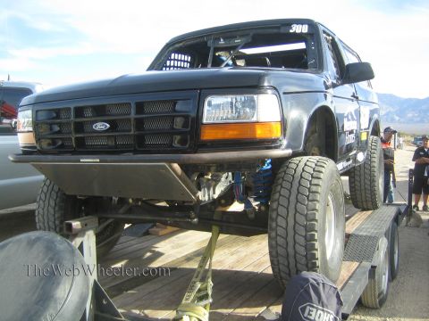 1990 Ford Bronco - Front view of a black Ford Bronco race truck numbered 300 showing its grille, lifted suspension and blue coilover shocks on a trailer.