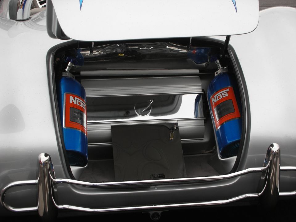 Silver car trunk open showing two blue NOS bottles, chrome trim, and a black square object