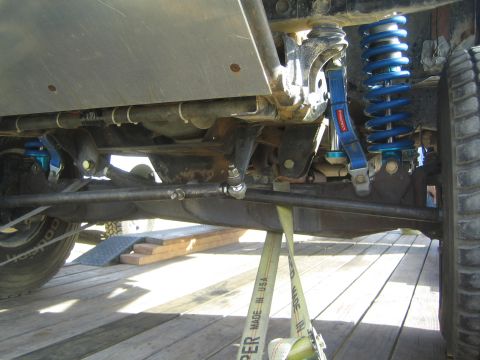 1990 Ford Bronco - Underbody detail of a lifted vehicle's front suspension showing a blue coilover shock, control arm and tie rod over wooden trailer planks.