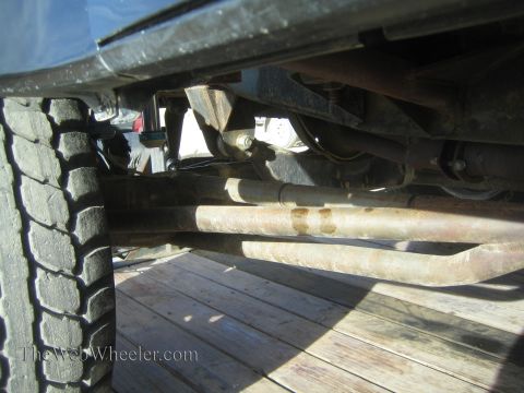 1990 Ford Bronco - Underbody view of a lifted vehicle showing twin radius arms, the tire tread and frame components above a wooden trailer deck.