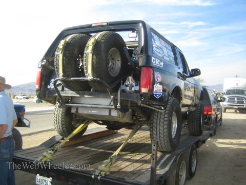 1990 Ford Bronco - Rear view of a black off-road race SUV carrying two spare tires on a swing rack, strapped to a flatbed trailer in a staging area.