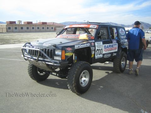 1990 Ford Bronco - black Jeep Cherokee race SUV numbered 1700 with a front tube bumper and many sponsor decals sits in a paved lot as a man walks past.