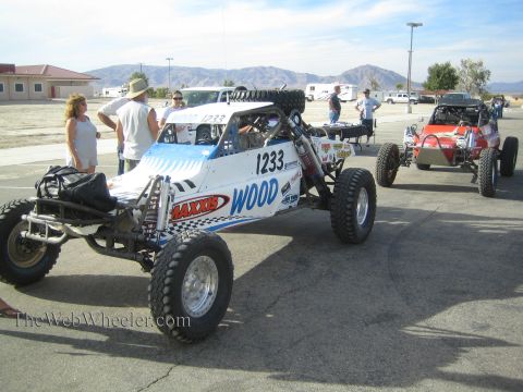 1990 Ford Bronco - white number 1233 Wood off-road race buggy and a red buggy sit in a paved staging lot with people and mountains in the background.
