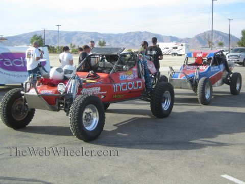 1990 Ford Bronco - red Ingold Motorsports off-road buggy and a blue buggy sit in a paved staging lot with spectators and desert mountains behind.