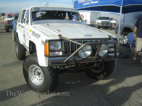 1990 Ford Bronco - white Jeep Cherokee race SUV numbered 1775 with a front tube bumper and round driving lights sits in a paved staging lot.