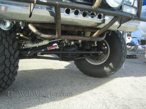 1990 Ford Bronco - Underbody close-up of an off-road vehicle showing the front solid axle, steering damper, tie rod and exhaust header tubes.